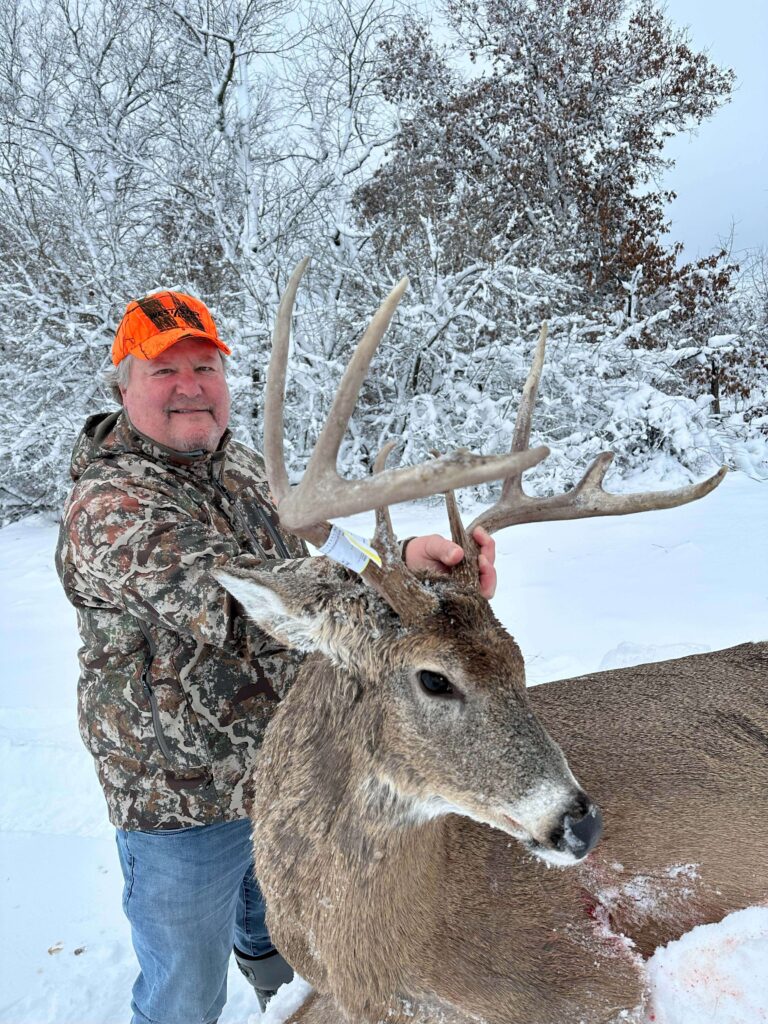 Man posing with deer, smiling with hat, in the snow.