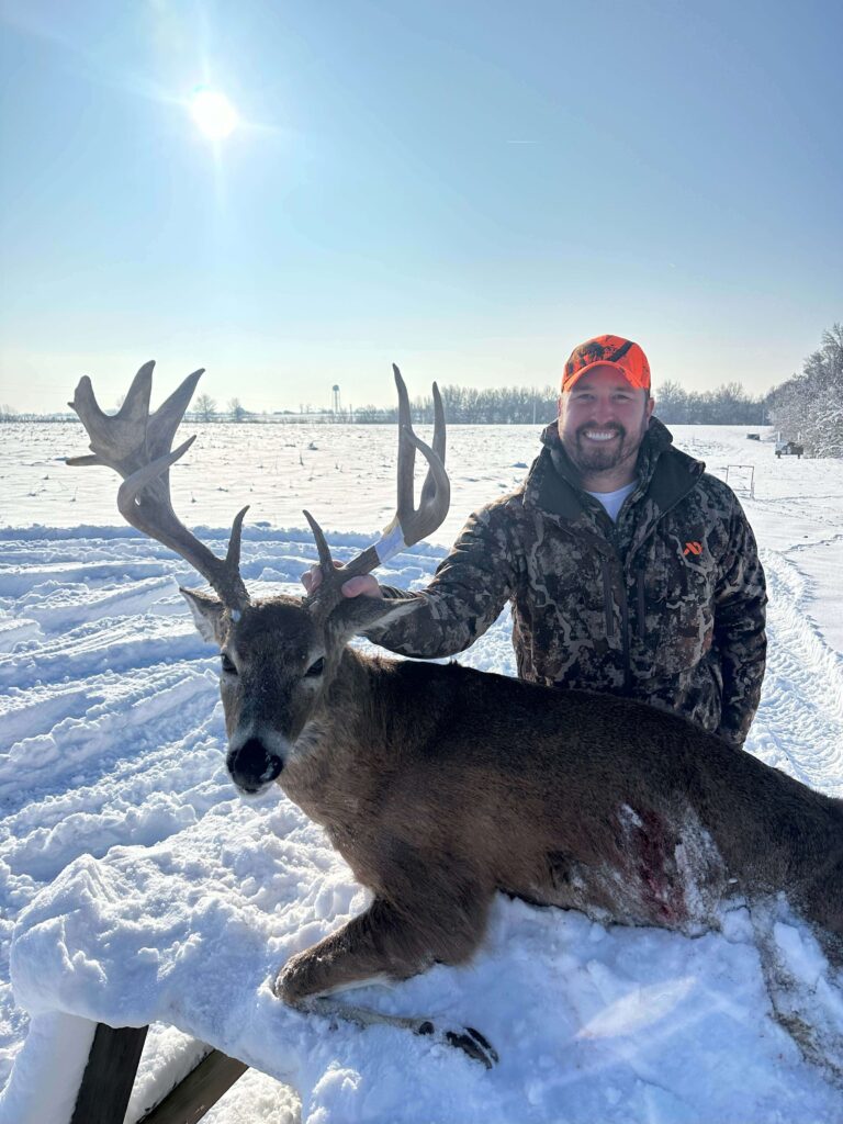 Man posing with deer, smiling with hat in the snow.