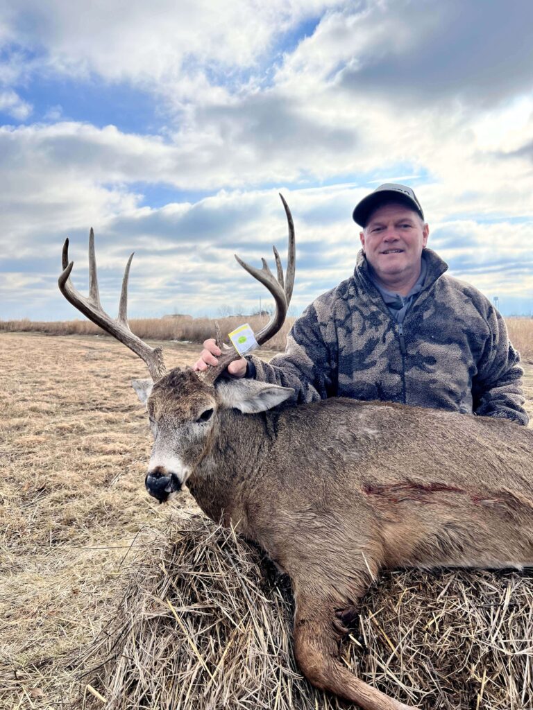 Man posing with deer, smiling with hat in the middle of a field.